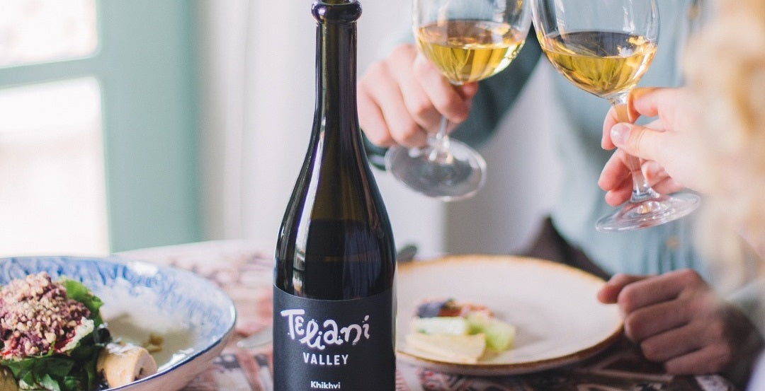 Teliani Valley, Winery 97 Khikhvi bottle on a dining table covered in a patterned table cloth. A dish of cheese and salad is is laid out on a bowl and two side plates and two people at the edge of the right of the frame, faces not visible, clink glasses.,
