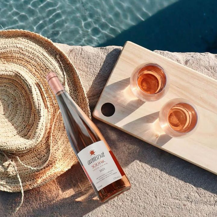 Bottle and glasses of rosé wine on a wooden board by a poolside with a straw hat.