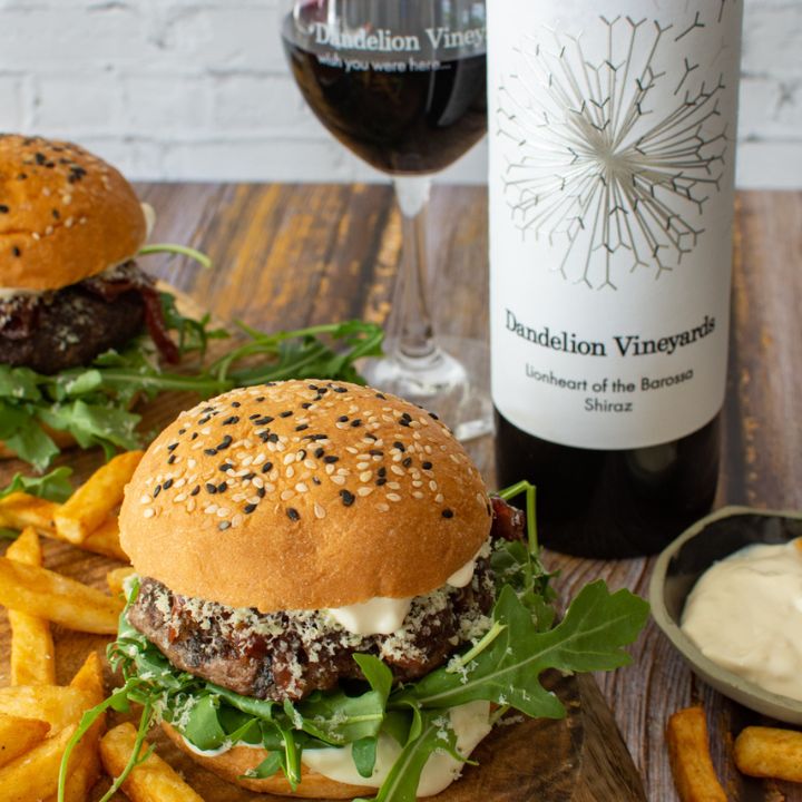Two burgers with fries, a glass of wine, and a bottle of Dandelion Vineyards Shiraz on a wooden table.