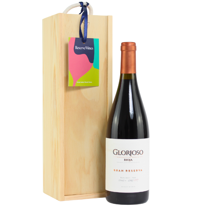 A bottle of Glorioso Rioja Gran Reserva 2017 red wine placed in front of its wooden gift box, with a colorful gift tag attached to the box.