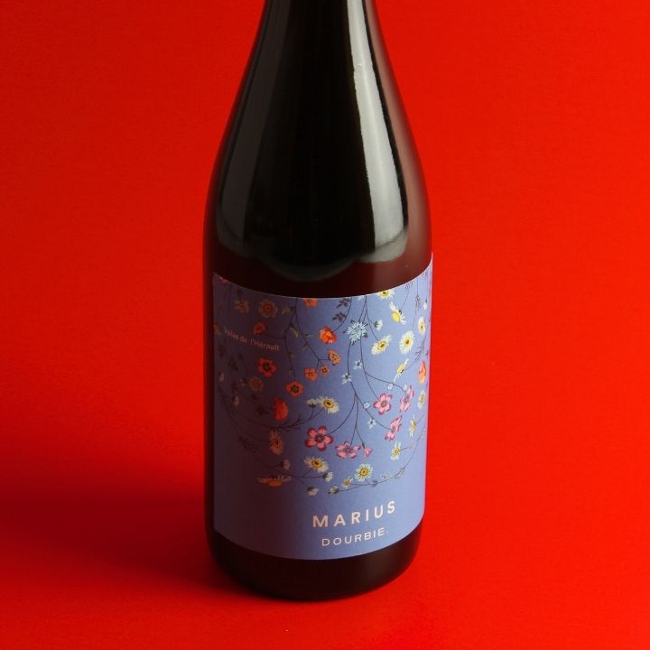 A bottle of La Dourbie, Marius Rouge 2023 wine with a light blue floral label, set against a solid red background.
