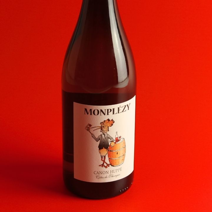 A dark brown glass wine bottle with a white label featuring the brand name 'MONPLEZY' and an illustration of a cartoon character beside a barrel, set against a solid red background.