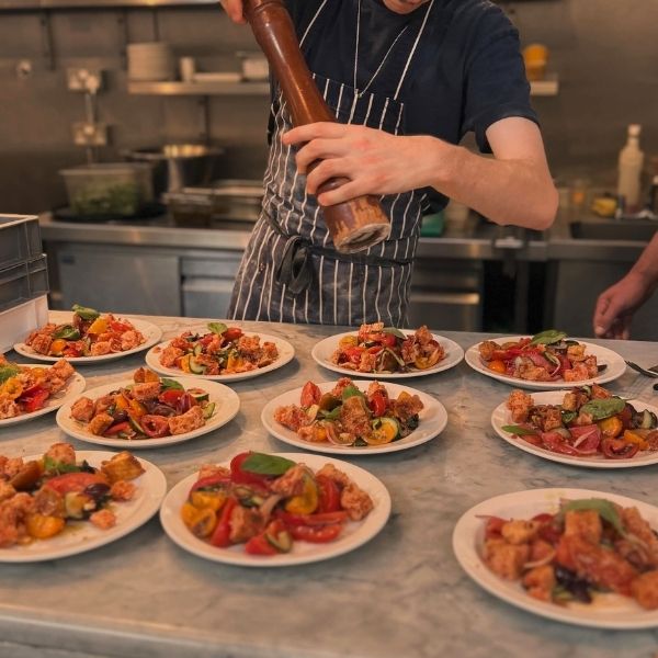 A chef in a kitchen grinds pepper over multiple plated seafood pasta dishes arranged on a marble counter.