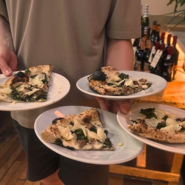 A person in a beige t-shirt holds four white plates, each with a slice of pizza topped with green spinach, white onions, and black olives, in a warm, dimly lit restaurant setting with wine bottles in the background.