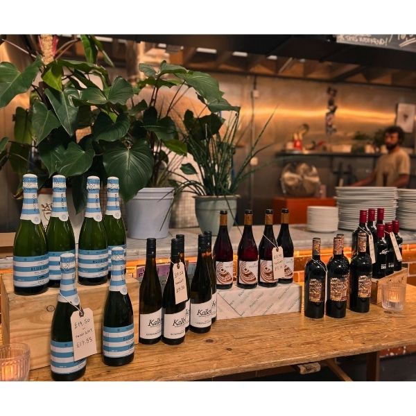 A variety of wine and sparkling beverage bottles arranged on a rustic wooden table in a warm, restaurant-like setting with potted plants and a blurred kitchen background.