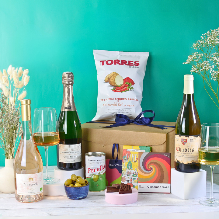 An assortment of gourmet food and wine presented in a gift hamper, including bottles of Chateau L'Escarelle Les deux Anges Rosé, Domaine Fournillon Chablis, and Domaine Landreau Crémant de Loire Brut, along with Torres Smoked Paprika Crisps and Perelló Pitted Gordal Picante Olives, plus oat-based chocolate bars with caramel and cinnamon flavors.
